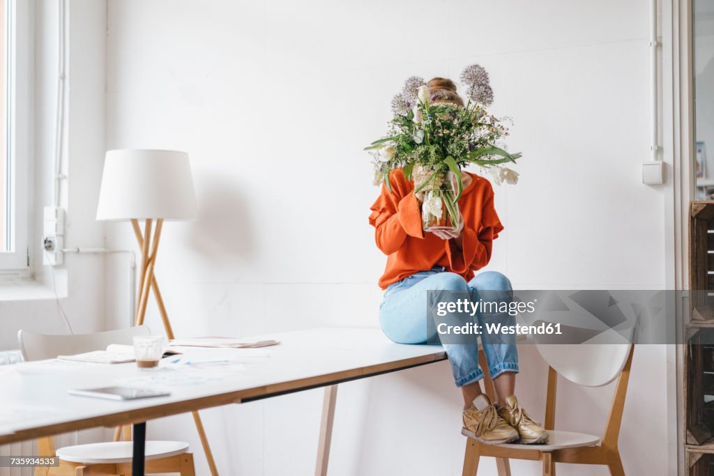 Young woman sitting on table holding flower vase in front of her face
