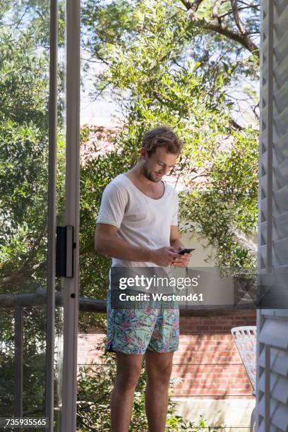 young man in swimming trunks and t-shirt using cell phone - zwembroek stockfoto's en -beelden