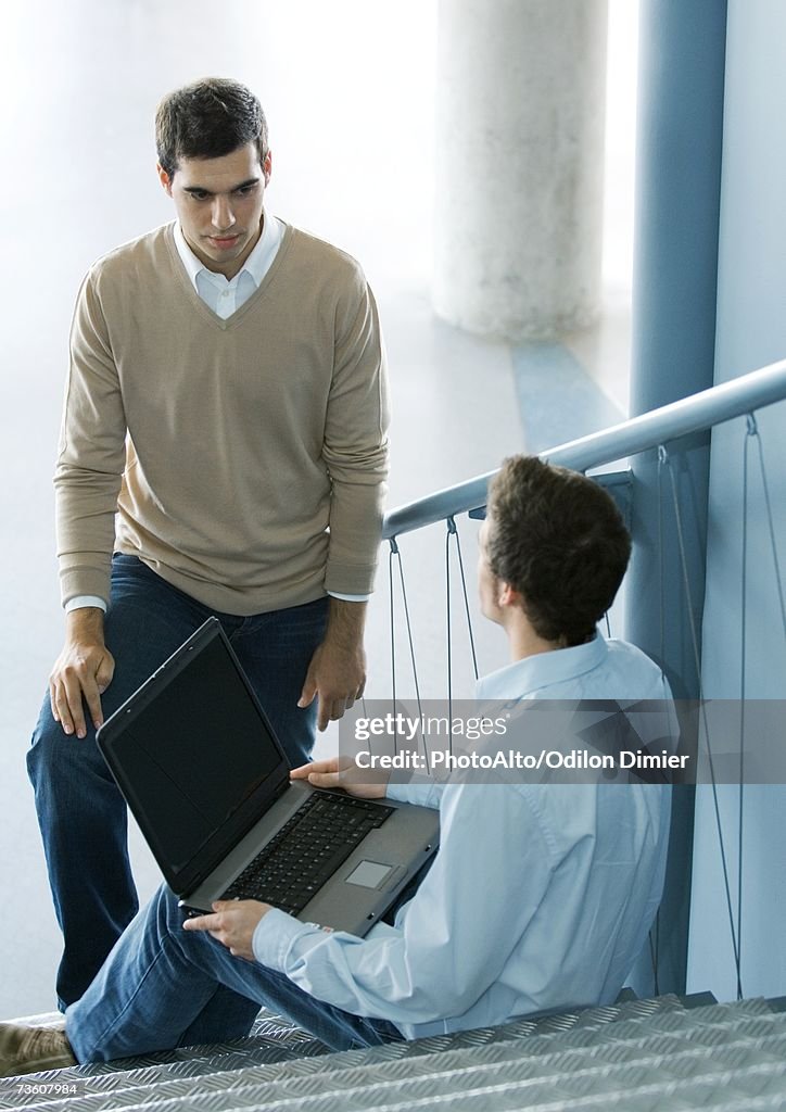 Two male colleagues talking, one sitting on stairs with laptop