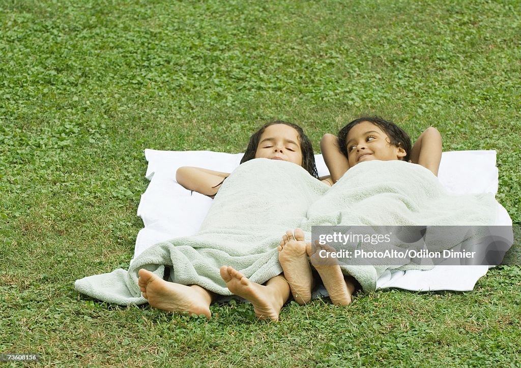 Two girls lying in grass, on a towel and covered with a towel