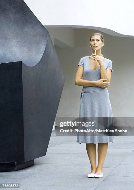 woman standing next to sculpture, holding pen to chin, full length - man standing full body stock-fotos und bilder