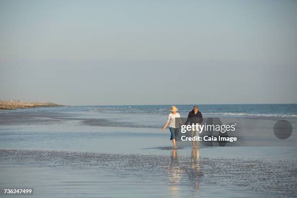 mature couple holding hands walking in sunny ocean beach surf - camber sands stockfoto's en -beelden
