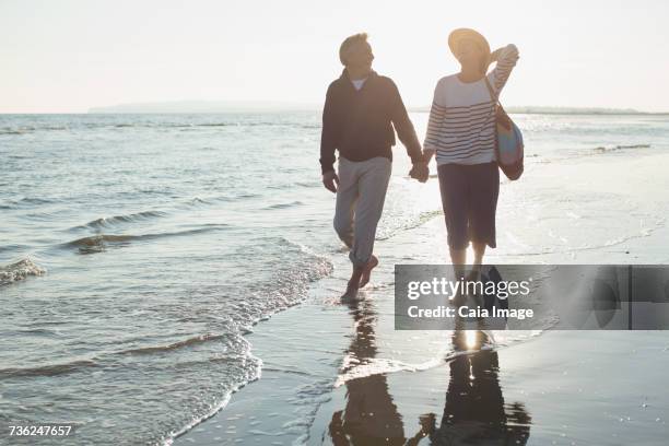 affectionate mature couple holding hands and walking on sunset ocean beach surf - camber sands ストックフォトと画像