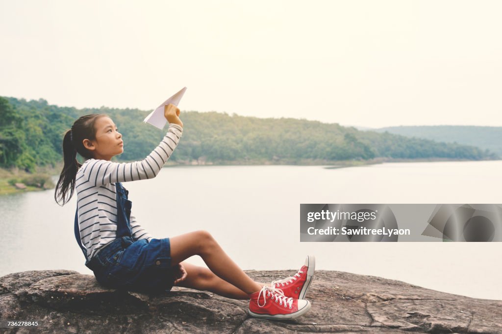 Side View Of Girl Playing With Paper Airplane While Sitting On Rock Against Lake