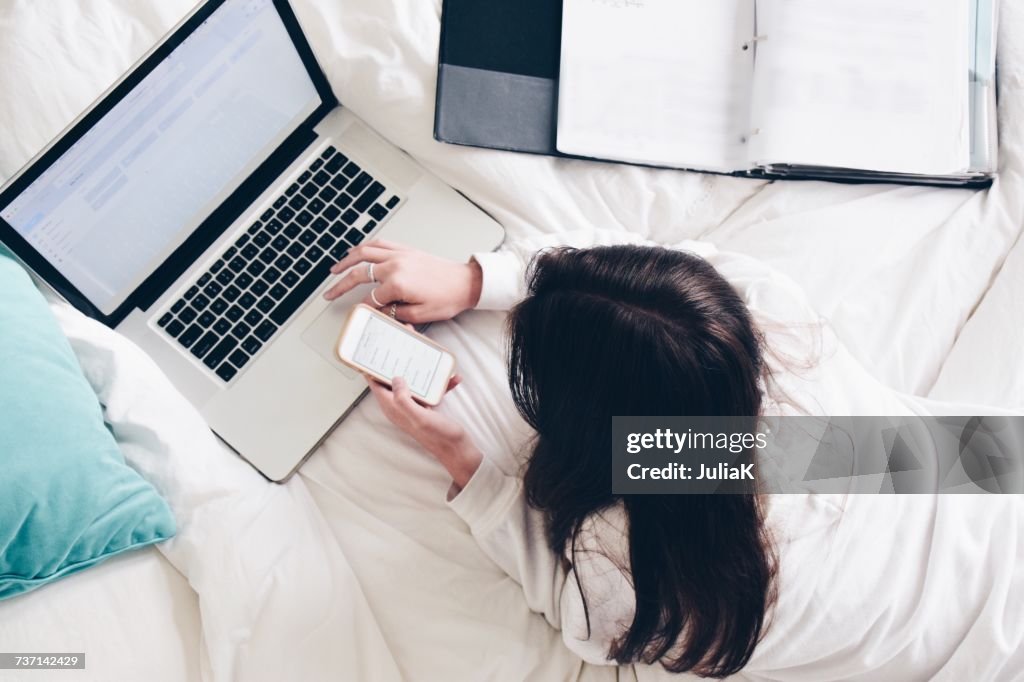 Teenage girl lying in bed using her laptop and mobile phone