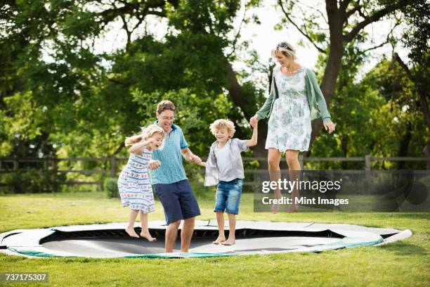 man, woman, boy and girl holding hands, jumping on a trampoline set into the ground in a garden. - trampoline jump stock-fotos und bilder