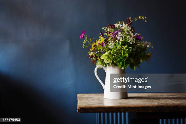close up of white jug with bunch of wild flowers on a wooden table, blue wall behind. - jug stock pictures, royalty-free photos & images
