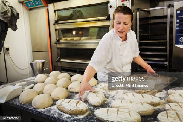 woman standing at a table, placing freshly baked loaves of bread on a wooden board. - baker stock-fotos und bilder