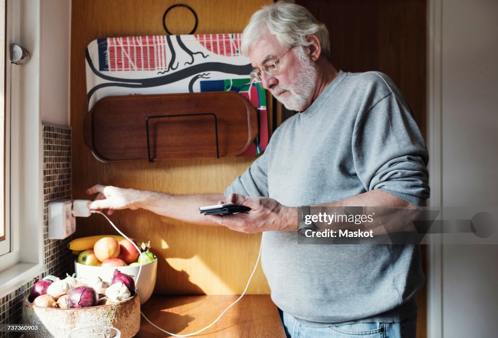 Senior man holding mobile phone while plugging charger to electrical outlet at domestic kitchen