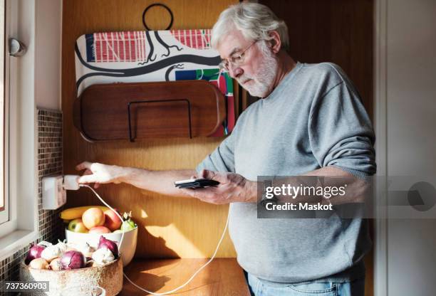 senior man holding mobile phone while plugging charger to electrical outlet at domestic kitchen - elkontakt bildbanksfoton och bilder