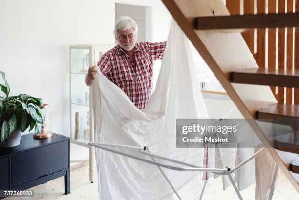 senior man folding white sheet for drying on rack in living room - bettbezug stock-fotos und bilder