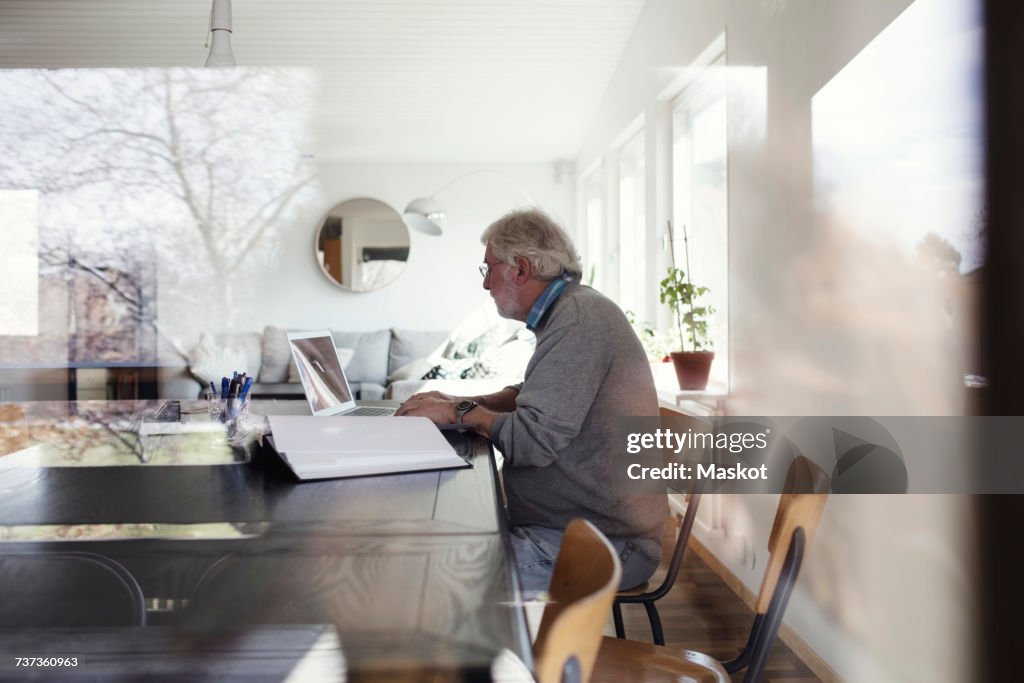 Senior man using laptop while sitting at table seen through window