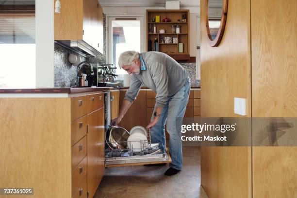 senior man putting plates in dishwasher at kitchen - witwer stock-fotos und bilder