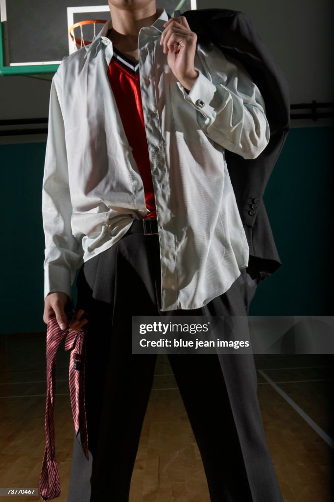 Young businessman poses holding his tie in one hand and his suit jacket thrown over his shoulder in a darkened gymnasium.