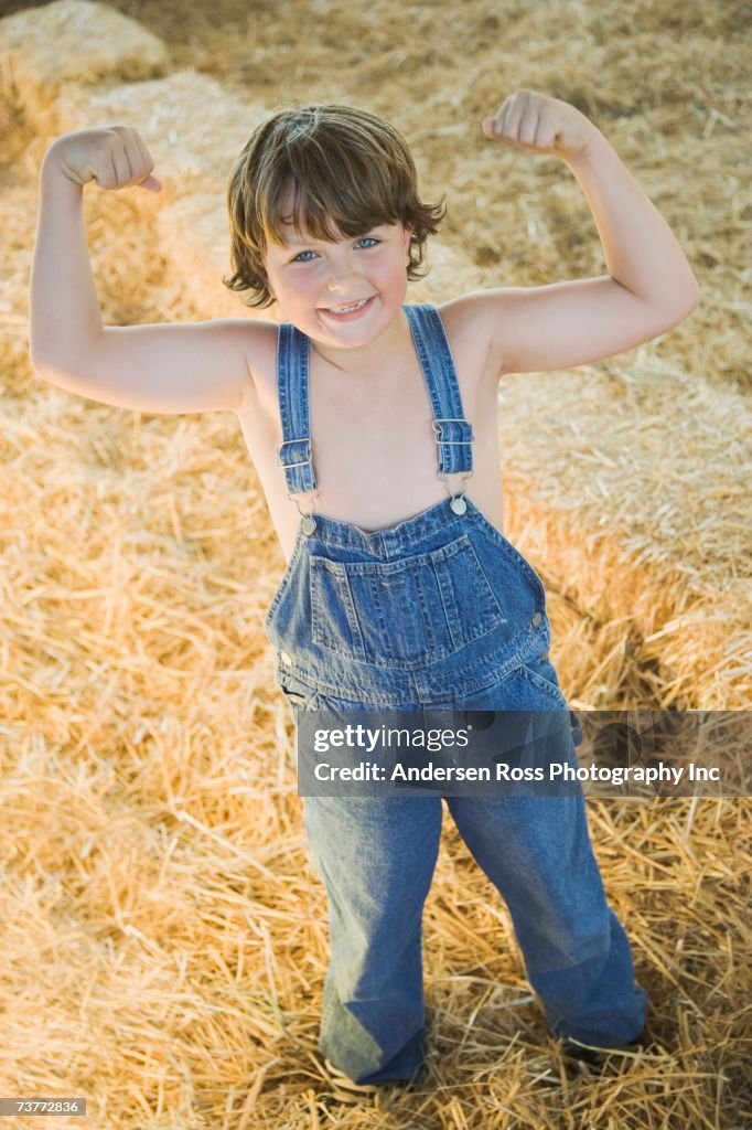 Young boy wearing overalls and flexing muscles in hay