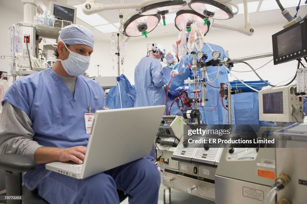 Doctor using laptop in operating room with surgery in background