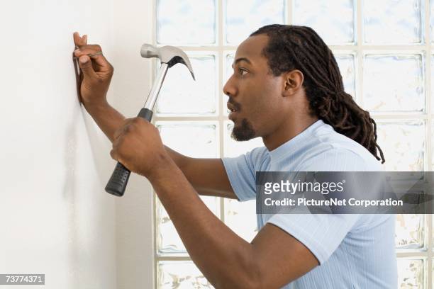 african man hammering nail into wall - hammer and nail fotografías e imágenes de stock