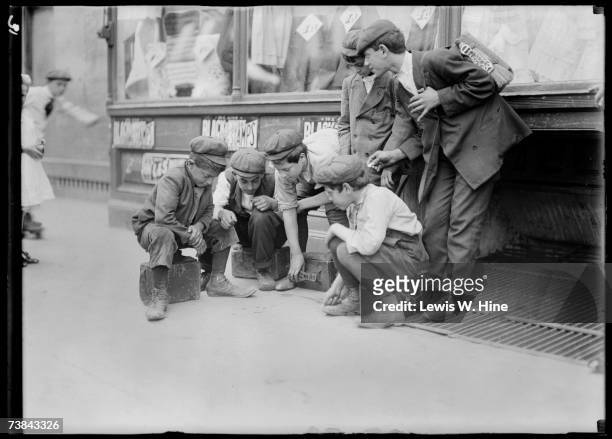 Group of newsboys and shoeshine boys in caps playing craps on a sidewalk in front of a store, entitled 'Newsies and Bootblacks Shooting Crap,' from...