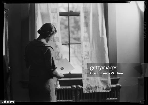 Middle-aged woman looks at a piece of paper with a leaf print on it as she stands at a curtained window by a radiator in a classroom, USA, early 20th...