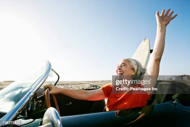 older caucasian woman in convertible car with surfboard on beach - auto convertibile photos et images de collection