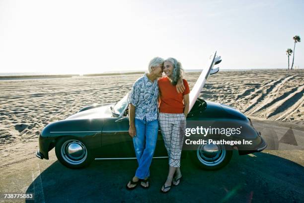 older caucasian couple leaning on convertible car with surfboard on beach - couple on the beach with car stockfoto's en -beelden