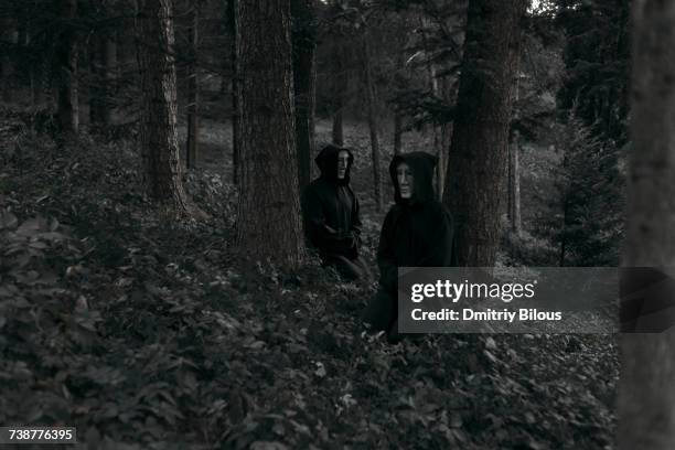 people wearing black robes and white masks sitting in forest - túnica fotografías e imágenes de stock