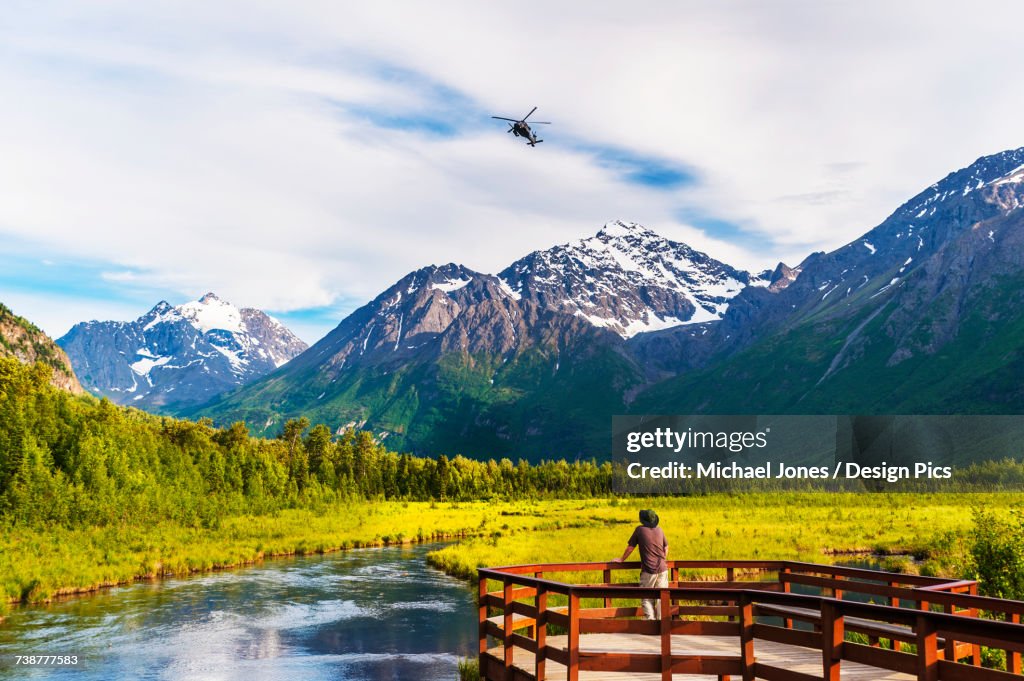A man is standing on the Eagle River Nature Center boardwalk while a Black Hawk helicopter flies over head in the Chugach State Park in Southcentral Alaska, USA