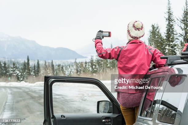 caucasian woman standing in car in winter photographing scenic view - auto von hinten winter stock-fotos und bilder