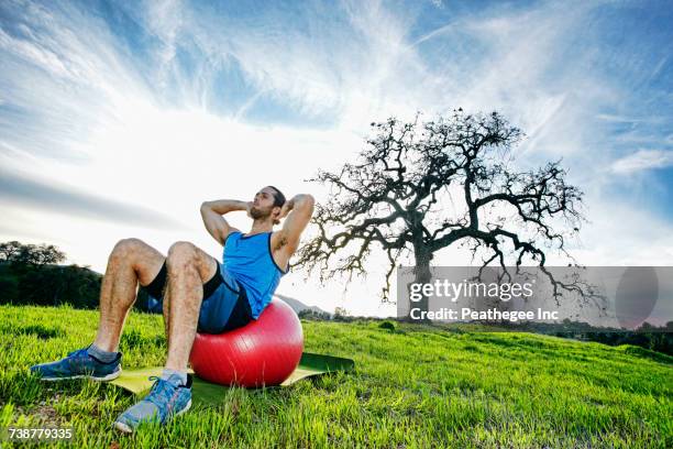 caucasian man doing sit-ups on fitness ball in field near tree - yoga ball fotografías e imágenes de stock