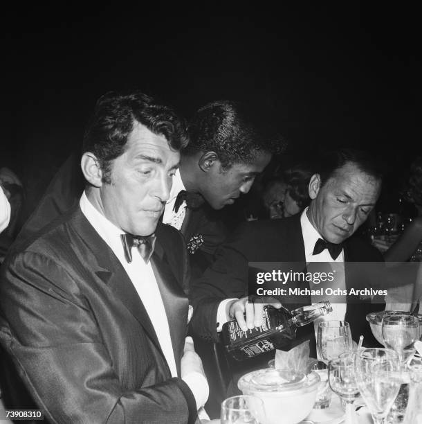 Singer Dean Martin and Sammy Davis, Jr. Sit at a table as fellow singer and Rat Pack member Frank Sinatra pours Jack Daniels from a bottle at the...