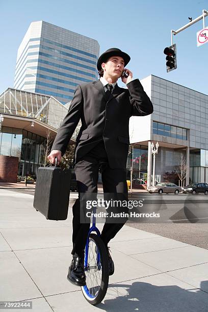 photo of a young businessman carrying a briefcase and talking on his cell phone while riding a unicycle - unicycle stock pictures, royalty-free photos & images