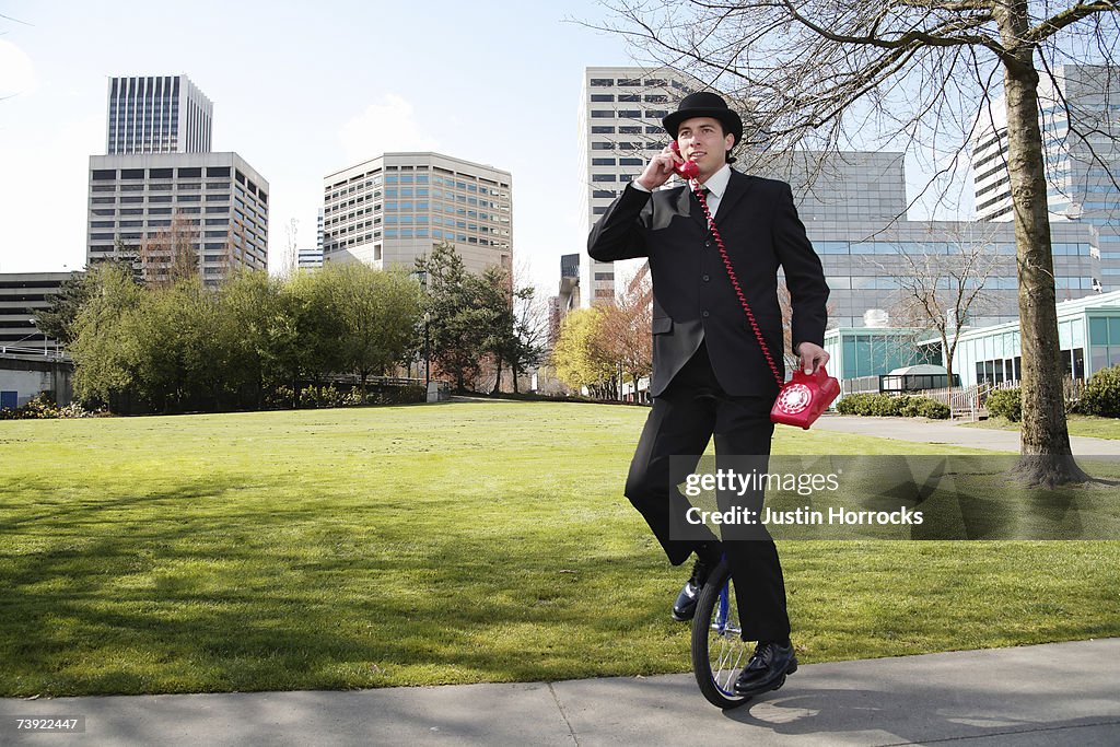 Photo of a young businessman talking on an antiquated red phone while riding a unicycle