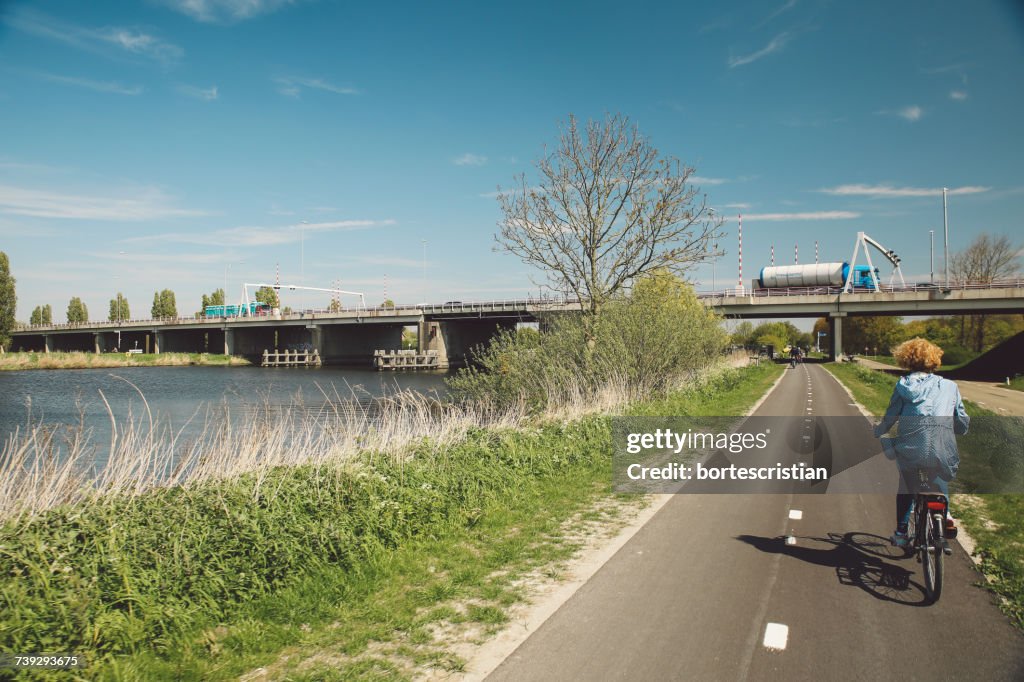 Rear View Of Woman Riding Bicycle On Road By River Against Sky