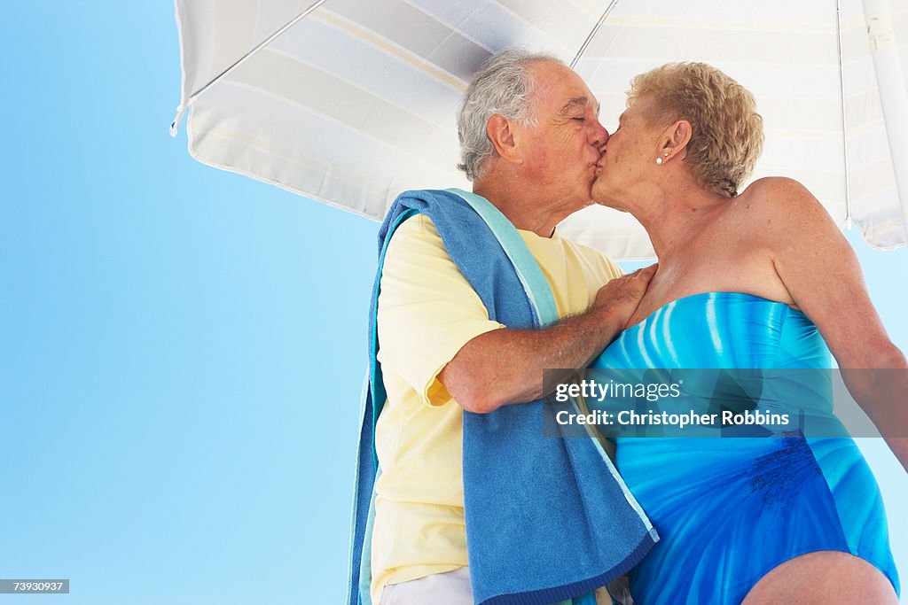 Senior couple kissing under beach umbrella, three quarter length