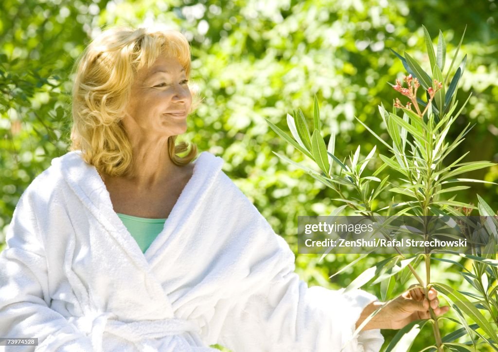 Senior woman in bathrobe smiling and examining branch of shrub