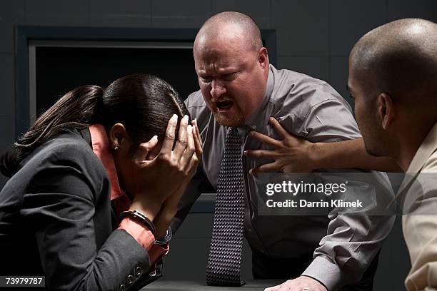 two men and handcuffed woman at desk in interrogation room, man shouting at woman - interrogation room stock-fotos und bilder