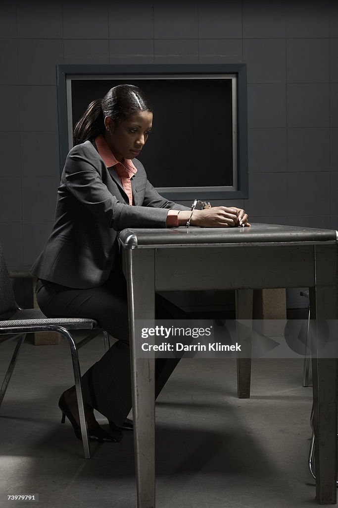 Young handcuffed woman sitting at table in interrogation room, side view