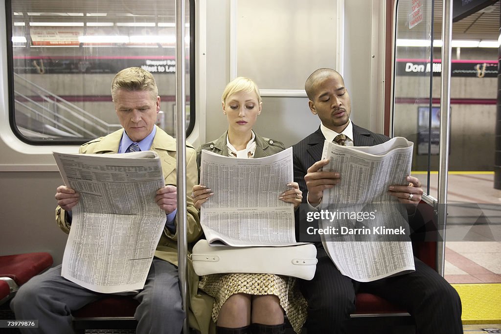 Two men and woman sitting in subway train side by side, reading newspaper