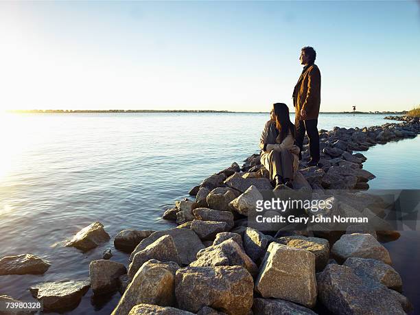 mature couple watching sunrise on rock breakwater - groyne photos et images de collection