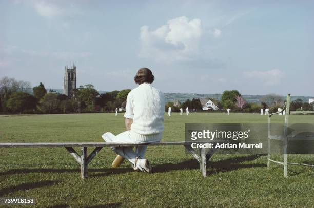 Player for the Blagdon Village 2nd XI cricket team sitting on a bench waiting to bat looks over to the game and St Andrew's Church on 20 May 1979 at...