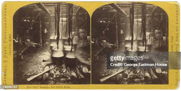 Stereoscopic view of several women and children as they pose with a number of steaming metal pots suspended over a fire on stripped tree branches,...