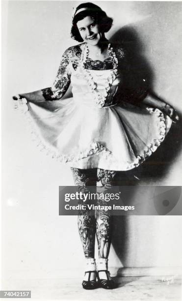 Portrait of an unidentified, heavily tattooed woman as she flairs the skirt of her dress and smiles, early twentieth century.