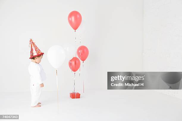 baby girl (15-18 months) wearing party hat standing in studio, looking at balloons and present - one baby girl only fotografías e imágenes de stock
