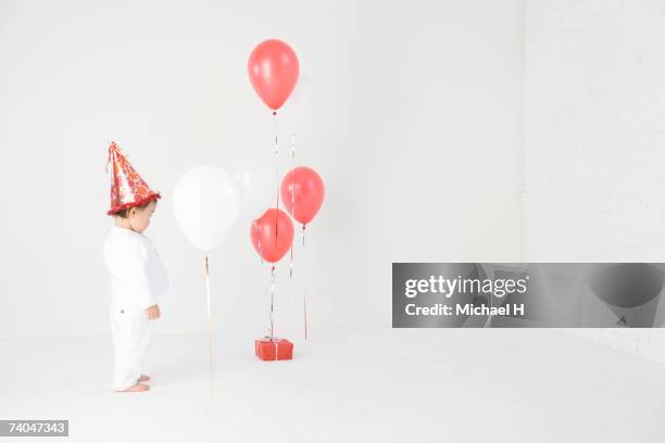 baby girl (15-18 months) wearing party hat standing in studio, looking at balloons and present - one baby girl only foto e immagini stock