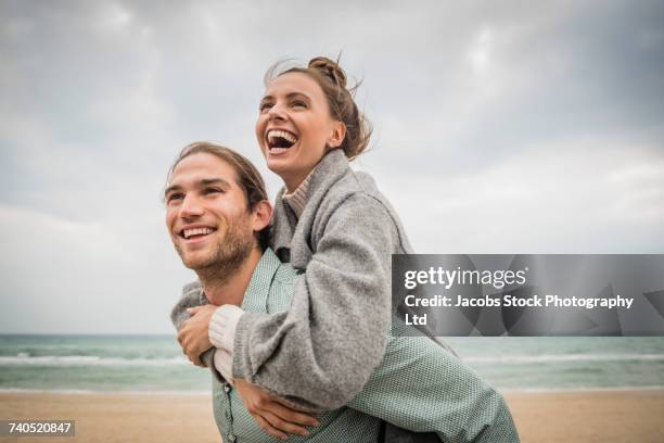 caucasian man carrying woman piggyback on beach - boyfriend stockfoto's en -beelden