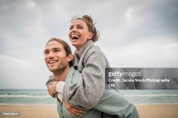 caucasian man carrying woman piggyback on beach - piggyback stock pictures, royalty-free photos & images