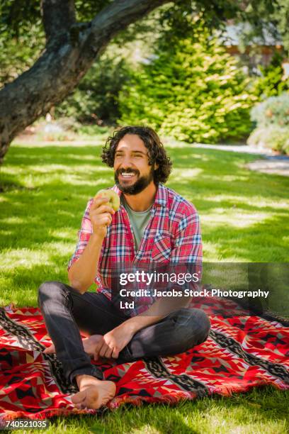 hispanic man sitting on blanket in grass eating fresh apple - picknickdecke stock-fotos und bilder