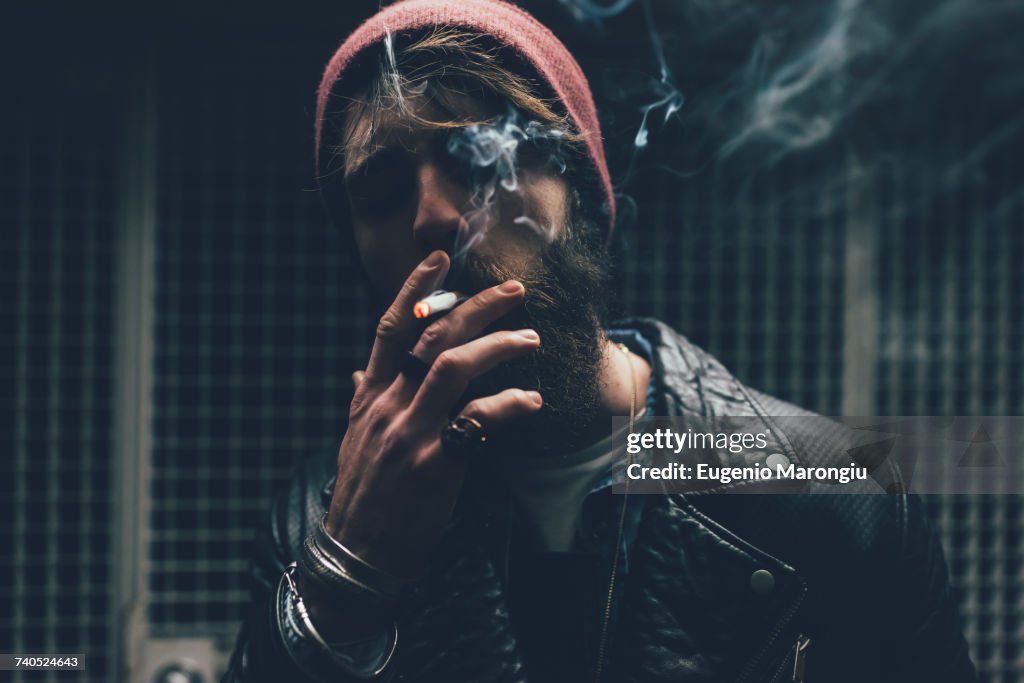 Young male hipster smoking cigarette in dark city doorway at night