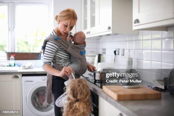 woman cooking in kitchen, baby strapped to body in sling, daughter standing beside her - busy mom stock-fotos und bilder