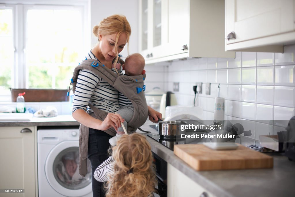 Woman cooking in kitchen, baby strapped to body in sling, daughter standing beside her